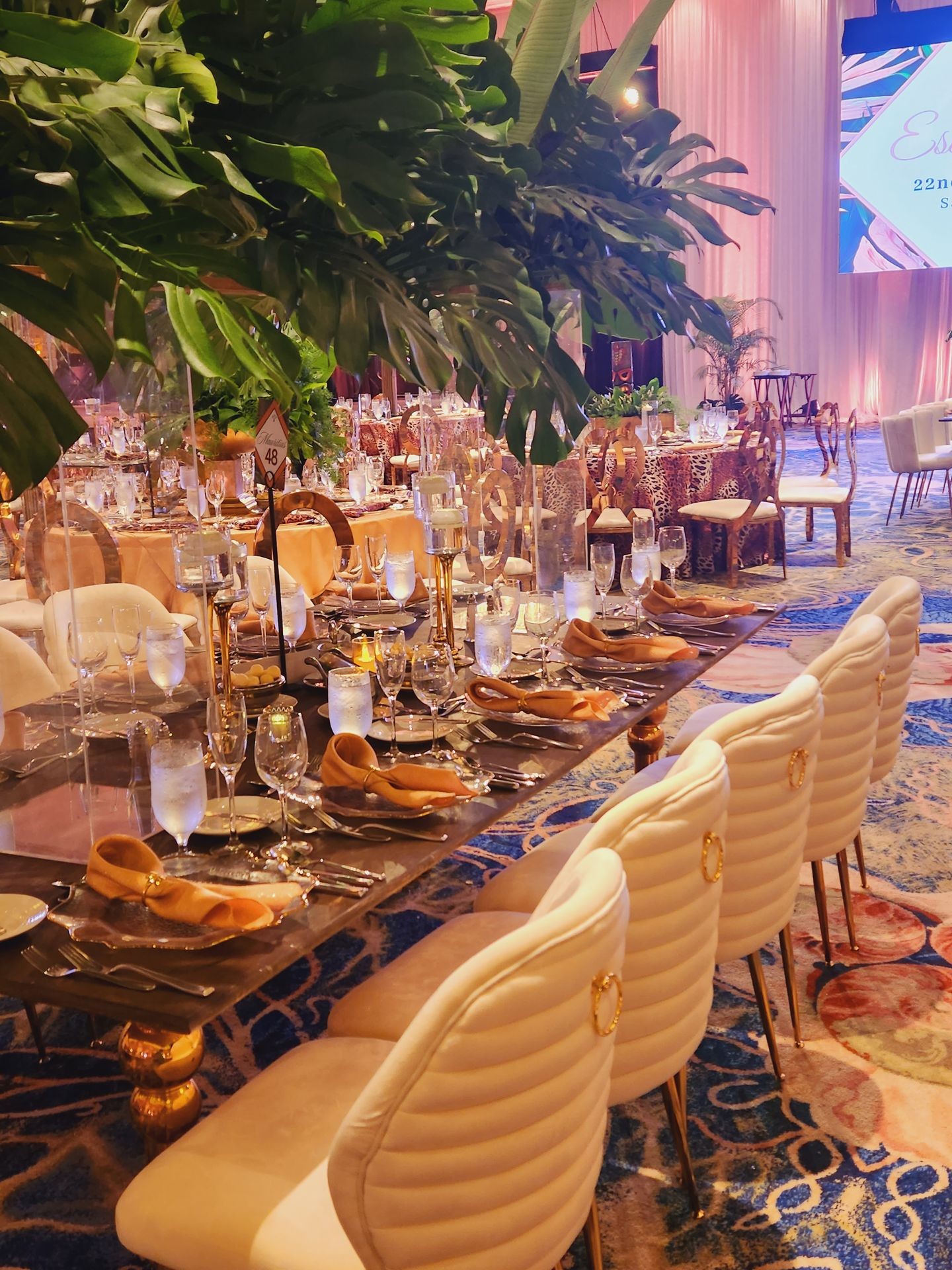 Elegant dining setup with white chairs, gold accents, and tropical greenery in a luxurious venue.