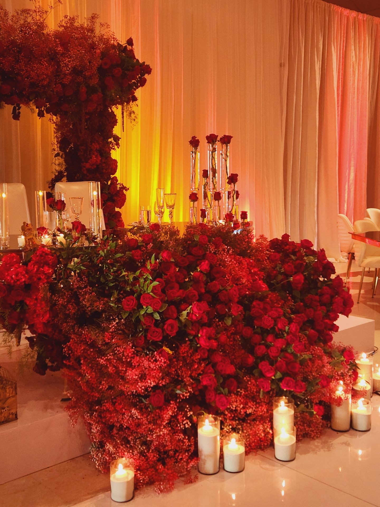 Elegant wedding decor with red roses, candles, and soft lighting against draped fabric backdrop.