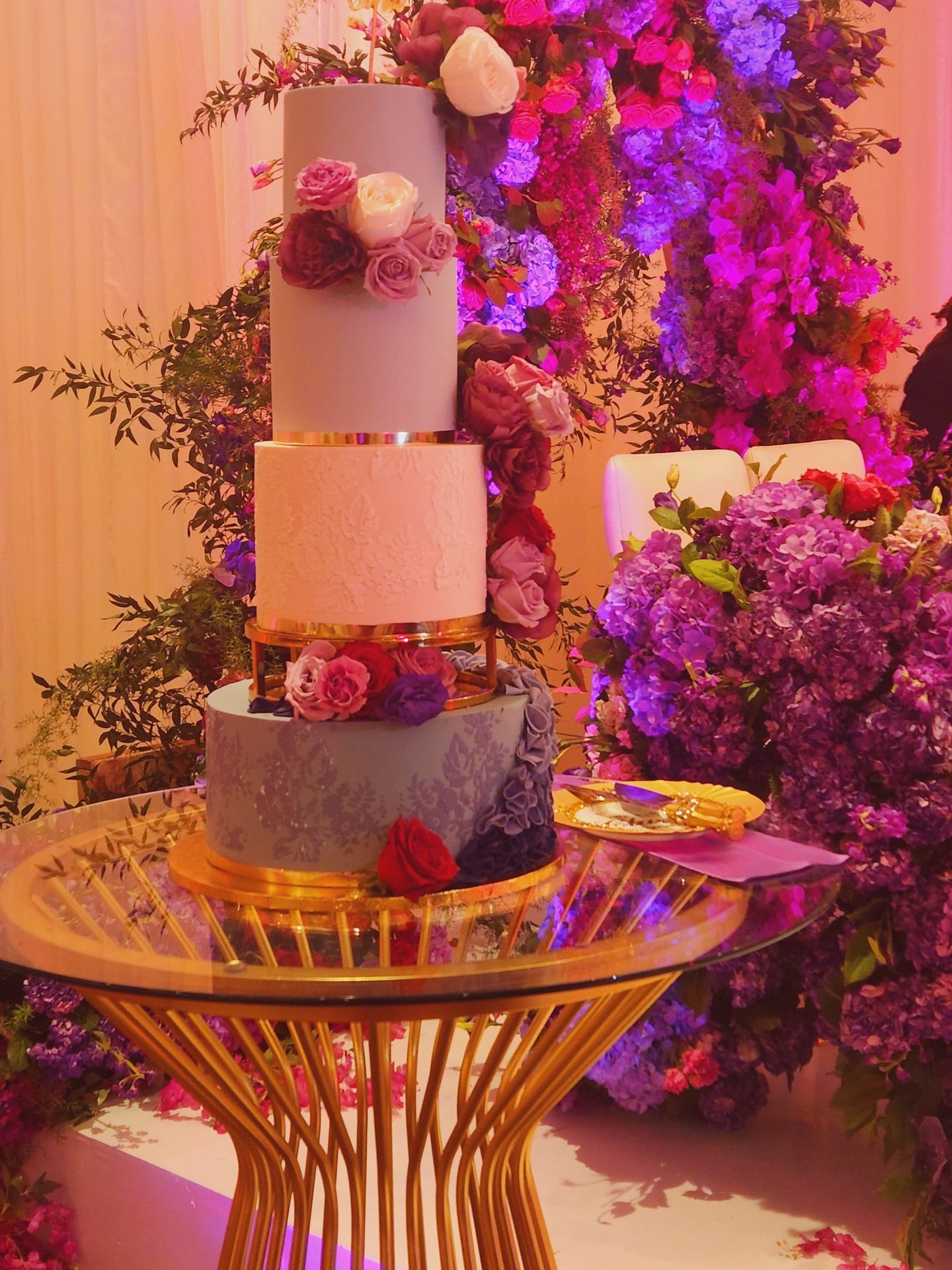 Elegant three-tiered cake adorned with purple and pink flowers on a glass table.