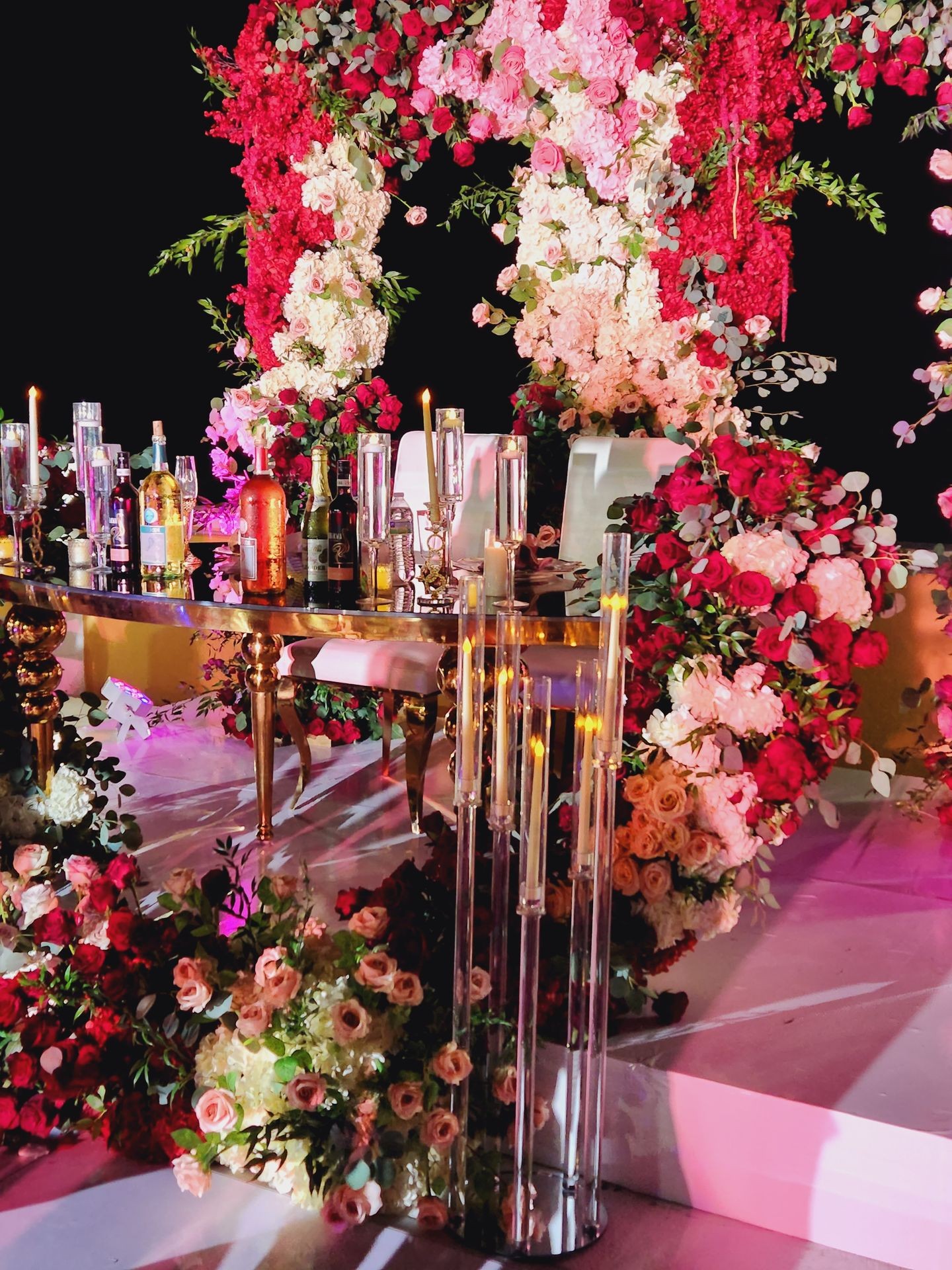 Elegant wedding setup with a floral arch, candles, and bottles on a reflective table.